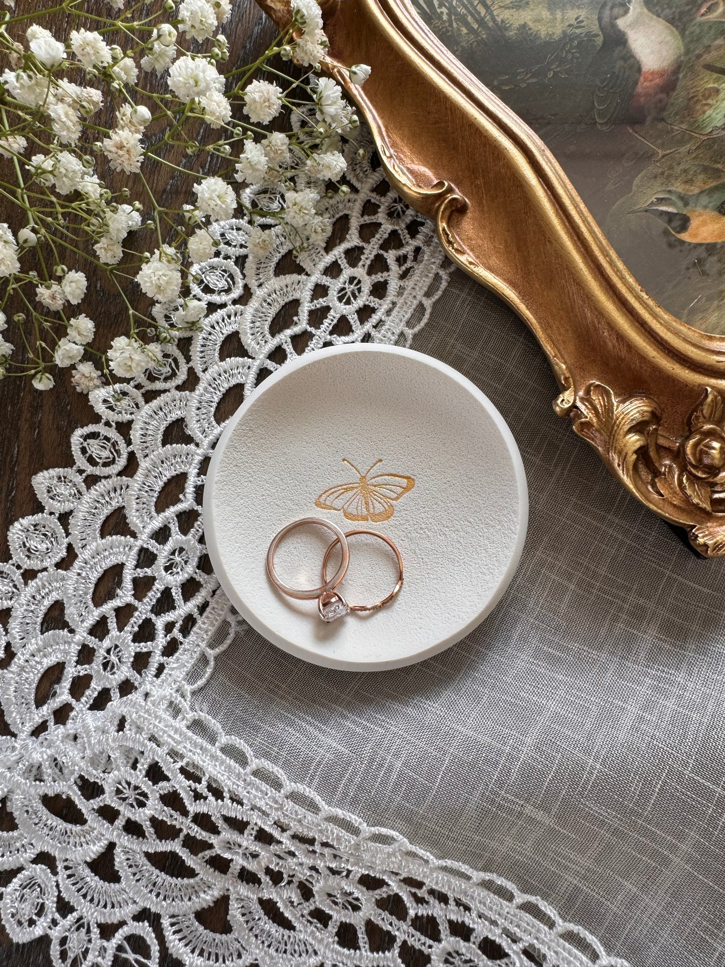 white clay ring dish with gold butterfly design in center in flat lay photo with babies breath and gold frame